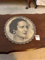 Top view of round picture frame with scalloped edges on wooden table showing black and white portrait of a woman with pearl earrings and necklace.