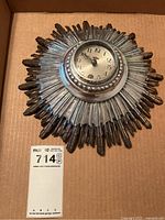 Top-down view of ornate sunburst clock front in cardboard box with lot tag