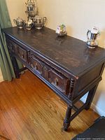Side view of antique solid wood buffet showing three drawers with ring pull handles and carved panels, standing on turned legs.