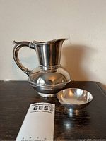 Silverplated jug and small bowl placed on a dark wood table showing signature and dimensions tag.