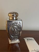 Full view of silver tea caddy resting on table next to auction tag