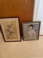 Two framed prints on the floor against a wood door, one black and white anatomical art and one Renoir reproduction portrait.