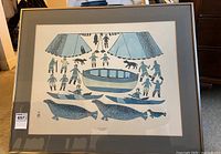Framed art print showing a hunting scene with Inuit figures, kayaks, animals, teepees in blue and black ink, with mat and frame visible.