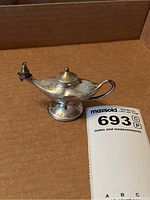 Miniature silver oil lamp resembling Aladdin's lamp shown from side angle with visible spout, lid, handle, and base.