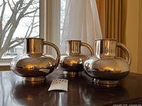 Three reflective silver-tone pitchers arranged on a table in front of a window