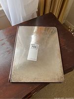 Closed silver-tone large guest book or menu cover lying on wooden table showing tarnished surface and minor scratches.