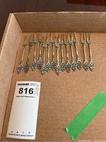 14 antique silver pickle forks laid out in a cardboard box, showing details of ornate handles and patina.