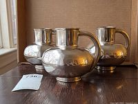 Three silver-tone pitchers on wooden surface in front of cardboard backdrop, lot tag visible