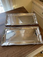 Two matching rectangular silver-tone trays with beaded rope edge on wood table