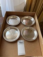 Four matching small silver-tone serving plates placed in a cardboard box, top view showing surfaces and rims with visible wear.