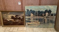 Two framed pastoral scene paintings side by side on floor against wall. One depicts trees and houses under cloudy sky; the other shows boats docked on water by a bridge.
