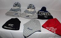 Photo of six tuques including two Royals knit with pom-poms, Canada knit with pom-pom, black Victoria fleece, striped Victoire knit, and red checkered knit