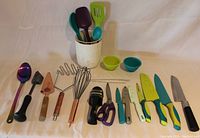 Photo of various kitchen utensils arranged on white cloth, showing spoons, spatulas, whisk, tongs, knives, scissors and measuring cups