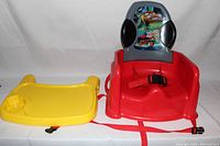 Red booster seat with grey backrest showing Lightning McQueen and Disney Cars graphic, black side pads, red harness, and separate yellow tray with cup holder placed beside it.