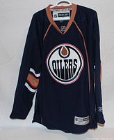 Front view of Edmonton Oilers Reebok NHL hockey jersey on hanger showing large team logo and tan trimmed collar.