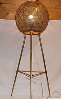 Side view of gold-tone metal tripod floor lamp with perforated spherical shade propped on tripod legs against wooden wall