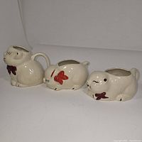Three ceramic animal-shaped vessels including two cats and one pig from side view, showing the red bows and cream color glaze.