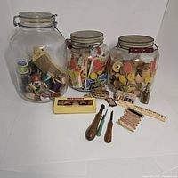 Glass jars filled with assorted sewing supplies including colored thread spools, needles in vintage packaging, and sewing tools.