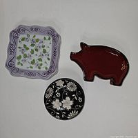 Top view of three plates: one square with purple floral pattern, one small round black floral plate, one red pig-shaped plate.