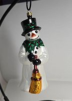 Front view of the snowman ornament showing walking snowman with black top hat, holly scarf, and broom