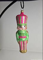 Front view of pink and green toy soldier Christmas ornament showing facial details, uniform and hat with striped pattern