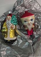 Two fragile Christopher Radko Christmas glass ornaments on gray padding showing detailed front features of both Santas.