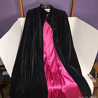 Black velvet cape hanging on a hanger showing opened front with vibrant hot pink satin lining inside.