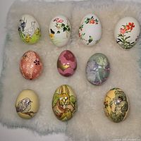 Photo of 10 delicate hand painted decorative eggs with scenes of animals, floral and abstract patterns arranged on soft white fabric.