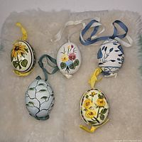 Top-down view of five hand painted eggshell hanging decorations on a soft white surface, showing sunflower, lily of the valley, multicolor flower basket, blue floral pattern, and yellow rose designs.