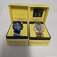 Two Invicta watches in their yellow presentation boxes, one blue-toned and one two-tone silver and gold, both with abalone faces.
