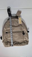 Front view of Michael Kors beige backpack with measurement tape showing approximately 14 inches in height, tags attached