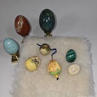 Seven miniature eggs displayed on a white fur-like surface: four natural stone eggs on brass stands, three smaller hand-painted decorative eggs. One reddish stone egg shows scratches.