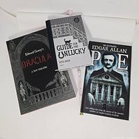 Three books arranged on flat surface showing covers of Dracula A Toy Theatre, Guide for the Unlucky, and Edgar Allan Poe pop-up books.