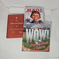 Photo showing three pop-up books including 'The Pop-Up Book of Nightmares', MAD political pop-up, and Sports Illustrated Kids pop-up book of sports.