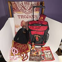 Photo showing maroon and gold USC Trojans throw blanket draped over chair, convertible rolling/backpack cooler in red and black with USC logos, needlepoint pillow cover, Marcus Allen bobblehead in front, and historic USC baseball program along with Sports Illustrated poster on the surface, plus red and pink beaded necklaces.