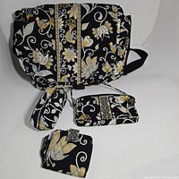 Photo showing the Vera Bradley messenger bag along with three matching accessories: wristlet, zippered pouch, and small wallet/cardholder, all featuring black background with yellow, white, and gray floral and bird patterns.