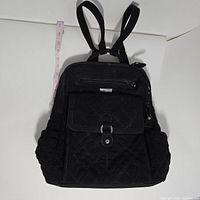Front view of black Vera Bradley quilted backpack showing adjustable shoulder straps, front flap pocket, side pockets, and measuring tape beside the bag to show size.