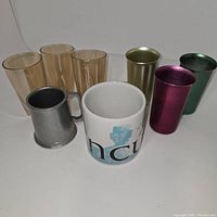 Eight drinkware items arranged on a plain surface: 4 amber glass tumblers, 3 metallic luster cups (gold, pink, green), 1 pewter-style mug, and 1 Starbucks ceramic mug.