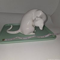 White porcelain cat figurine sitting with head bent down, shown on green tray with measuring tape indicating length of about 8 inches.