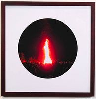 Framed digital photograph showing the full framed artwork with white matte and reddish-brown wooden frame. The photograph depicts a tall, bright bonfire at night with silhouettes of people and trees.