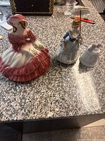 Group photo of three porcelain figurines on granite countertop; a woman in pink dress, another woman in white dress with parasol, and a small bell-shaped piece.