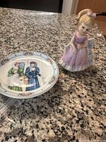 Photo showing porcelain figurine of a young girl in pink dress and a decorative plate with painted Victorian-style women on countertop.