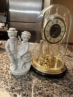 Porcelain figurine of a boy and girl in vintage clothing standing beside a glass dome clock with black and gold face.