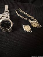 Three watches laid out on black fabric: one large wristwatch with silver metal band and black dial, one small bracelet watch with gold-tone band, and one small watch without band.