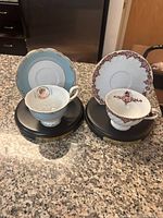 Two porcelain tea cup and saucer sets displayed on black presentation stands on a granite counter. One set is blue with gold and floral patterns, the other white with maroon ornate designs.