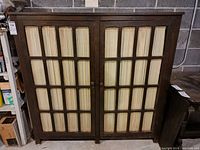 Front view photograph of antique wooden display cabinet with two glass paneled doors showing significant wear and scratches on the wood surfaces. The cabinet stands against a gray concrete wall.