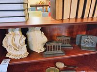 Photo showing white chalkware nautical ship bookends, brass Greek column bookends, and metal bookend with rider sculpture on wooden shelf.