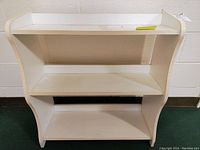 Side view of the white wooden bookshelf showing three shelves and curved panel edges with some dirt and scratches.