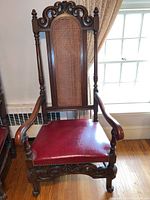 Full front view of the antique carved arm chair showing the leather-like red seat and caned backrest.