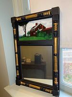 Full view of framed mirror with painted landscape panel on top and reflective mirror below, set on a window ledge.